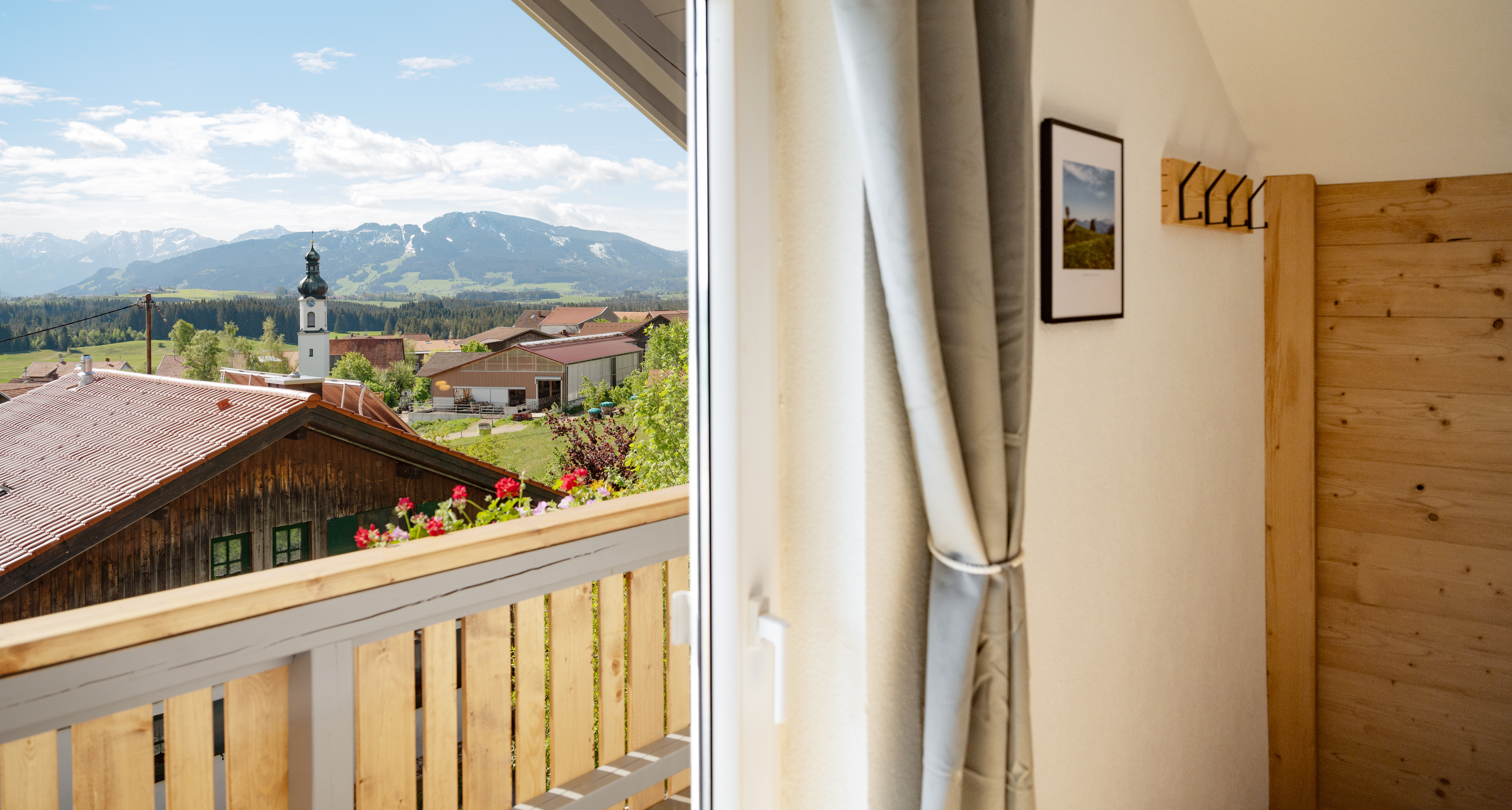 Blick vom Holzbalkon mit Geranien über den Bauernhof und den Kirchturm auf die Allgäuer Berge