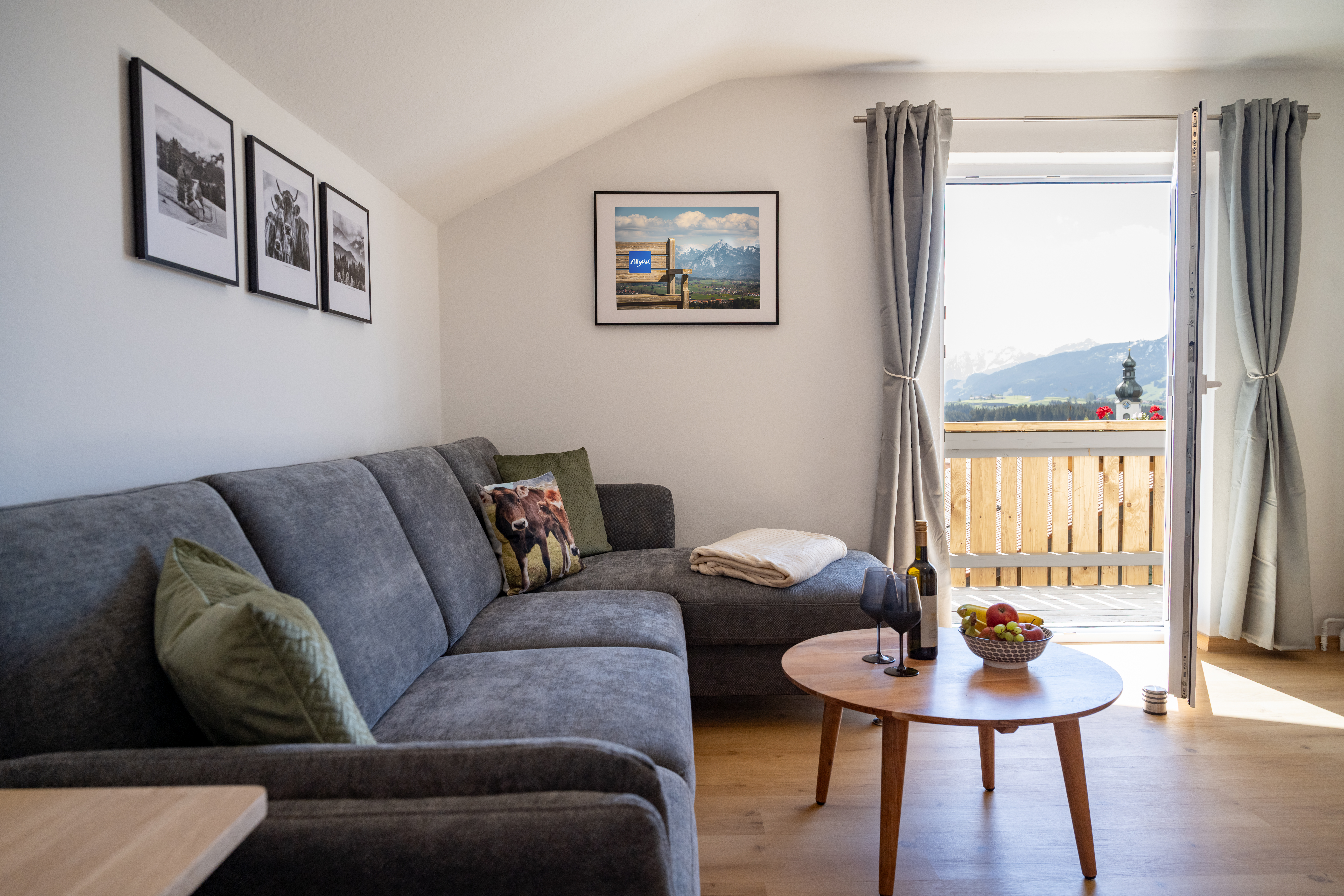Wohnzimmer mit grauem Ecksofa, rundem Holztisch und offener Balkontür mit Bergblick